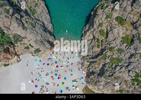 Blick aus der Vogelperspektive auf den Strand von Sa Calobra mit den Leuten, die im klaren blauen Wasser schwimmen und auf dem felsigen Sand liegen, mit Handtüchern und Sonnenschirmen zwischen zwei Bergen Stockfoto