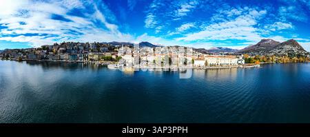 Blick aus der Vogelperspektive auf die malerische Hafenpromenade mit ruhigem See, umgeben von lebendiger Stadt und Bergen unter blauem Himmel, Lugano, Schweiz. Stockfoto