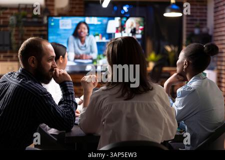 Geschäftsleute arbeiten bei nächtlichen Büromeetings zusammen. Eine Videokonferenz wird auf dem digitalen Monitor angezeigt, während Teammitglieder Marketingstrategien besprechen und Finanzdaten überprüfen. Stockfoto