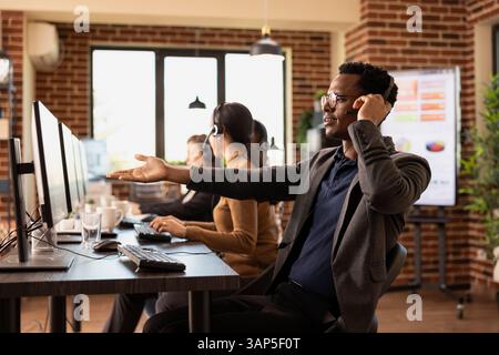 Afroamerikanischer Telemarketer mit Headset, der frustriert auf den Bildschirm zeigt und eingehende Kundenanfragen bearbeitet. Schwarzer Mann, der im Büro arbeitet, umgeben von Technologie und Kommunikationsgeräten. Stockfoto