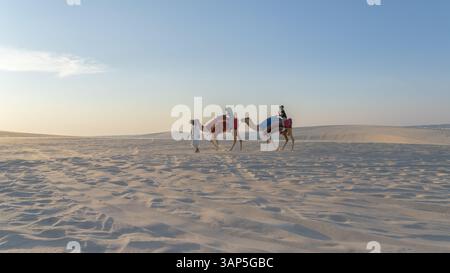 Doha, Katar, 24. Februar 2025: Eine Gruppe von Menschen, die auf Kamelen durch die goldenen Wüstendünen reiten, symbolisiert Ramadans spirituelle Reise und sein Erbe. Stockfoto