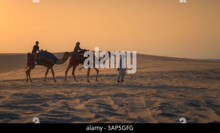 Doha, Katar, 24. Februar 2025: Eine Gruppe von Menschen, die auf Kamelen durch die goldenen Wüstendünen reiten, symbolisiert Ramadans spirituelle Reise und sein Erbe. Stockfoto