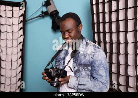 Professioneller männlicher Fotograf, der die Kamera hält und seine Bilder während eines Fotoshootings im Studio ansieht. Junger schwarzer Kameramann, der auf seinem dslr-Bildschirm steht und glücklich Bilder ansieht. Stockfoto