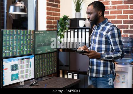 afroamerikanischer Investor, der mit seinem Tablet steht und Forex-Charts auf mehreren Bildschirmen überwacht. Das Büro verfügt über eine Backsteinmauer, die für Finanztechnologien gesorgt ist. Stockfoto