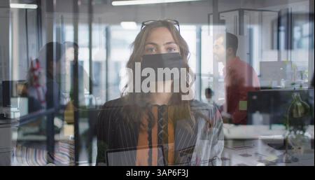 Komposit aus kaukasischer Frau, die im Büro eine Gesichtsmaske trägt, und Kollegen, die sprechen Stockfoto