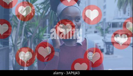 Bild von Social-Media-Herzsymbolen über lächelnder afroamerikanerin, die ihr Smartphone auf der Straße nutzt Stockfoto