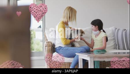 Bild von Herzen über kaukasierin und Tochter, die Gesichtsmasken tragen und mit Hund spielen Stockfoto