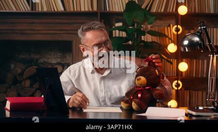 Glücklicher alter kaukasischer Mann Senior Großvater Geschenk Teddybär Bogen feiern Feiertag Gratulation Enkeltochter Enkelsohn sieht gerahmt Foto schreiben aus Stockfoto
