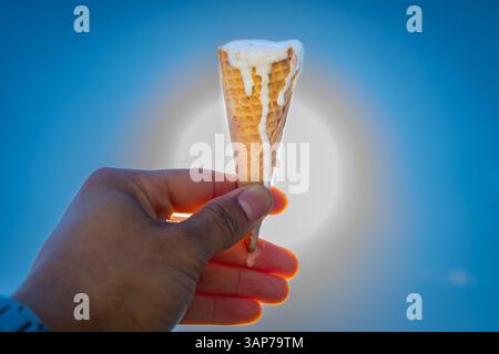Eine Hand, die einen Eiskegel mit einem weißen Niesel hält. Die Sonne scheint hell im Hintergrund und schafft eine warme und einladende Atmosphäre Stockfoto