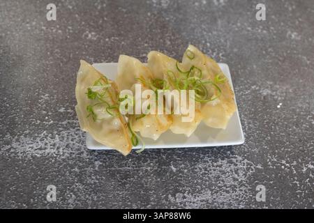 Vier köstliche, in der Pfanne gebratene Gyoza-Knödel, garniert mit gehackten grünen Zwiebeln, sitzen auf einem weißen Teller auf einer grauen Oberfläche Stockfoto