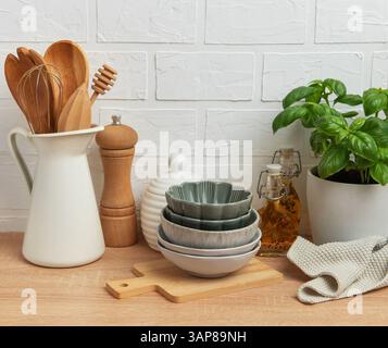 Verschiedene Küchenutensilien, darunter Schüsseln, Holzlöffel, Pfeffermühle, Ölflaschen, und eine Basilikumpflanze, die ordentlich auf einer hölzernen Arbeitsplatte angeordnet sind Stockfoto