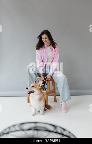 Eine modische junge Frau sitzt bequem in einem modernen Studio mit ihrem fröhlichen Corgi und schafft eine verspielte Atmosphäre, die ihre Gefährten zur Schau stellt Stockfoto