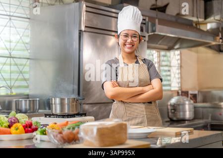 Porträt einer jungen Küchenchefin in einer professionellen Küche, die ihre Arme kreuzt und hinter einer Theke steht, gefüllt mit frischem Gemüse und gekocht Stockfoto