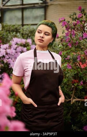 In einem Gewächshaus konzentriert sich eine selbstbewusste junge Frau mit grünen Haaren auf ihre Arbeit unter Blumen. Stockfoto