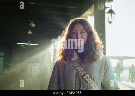 Eine junge Frau in einem warmen Wintermantel lächelt in einem Lichtstrahl auf dem rialto-Markt in venedig, italien, mit goldenem Sonnenlicht, das ihre lockigen Haare beleuchtet Stockfoto