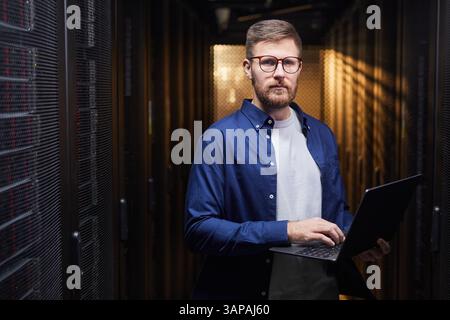 Porträt eines Mannes mit einer Brille, der im Serverraum steht und einen Laptop hält, während er Server verwaltet. Ein blaues Hemd tragen und die Serverleistung analysieren Stockfoto