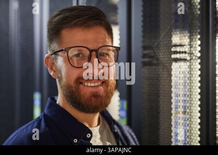 Porträt eines lächelnden bärtigen Mannes, der eine Brille trägt und im Serverraum steht, der voll mit Geräterücken und Kabeln ist, was ein selbstbewusstes und zugängliches Verhalten zeigt Stockfoto