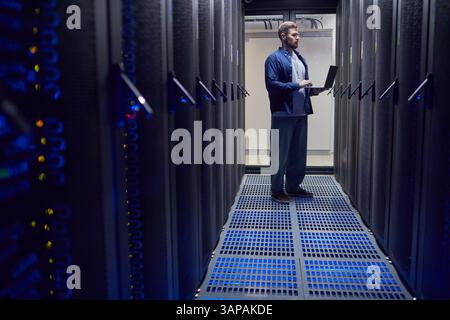 Männlicher Techniker, der im Rechenzentrum steht und einen Laptop zur Überwachung und Wartung der Serverleistung verwendet Stockfoto