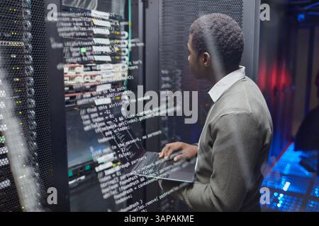Afroamerikanischer Techniker überprüft Datensysteme in modernen Rechenzentren, überwacht den Status auf Laptops zwischen Servern und Kabeln, um optimale Leistung und Sicherheit zu gewährleisten Stockfoto
