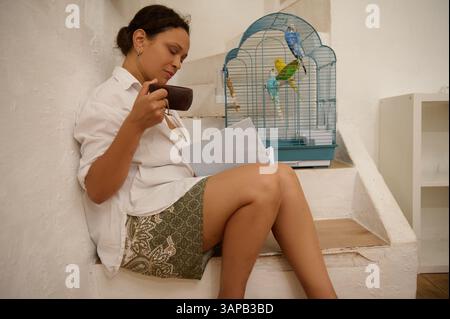 Eine Frau sitzt friedlich auf der Treppe, schlürft ein Getränk, liest und genießt die Gesellschaft farbenfroher Vögel in einem Käfig. Die Szene fängt Ruhe ein, Stockfoto