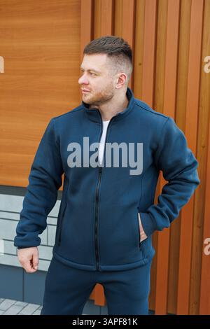 Ein selbstbewusster junger Mann, gekleidet in einer stilvollen marineblauen Jacke und modischen Hosen, posiert im Freien vor einem auffälligen Hintergrund des Contempo Stockfoto