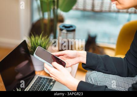Moderne Frau, die auf Smartphone in der Nähe von Laptop in gemütlichem Zuhause scrollt, Fernarbeitskonzept gelbes Sofa grüne Pflanzen Stockfoto