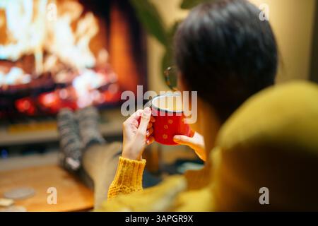 Frau genießt gemütlichen Winterabend mit heißem Getränk in der Hand, sitzt am Kamin, festliche Atmosphäre Stockfoto