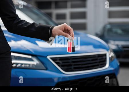 Hand mit Autotasten, selektives Fokusbild. Nahaufnahme der Seitenansicht der Frau, die Autoschlüssel übergibt, Vorderteil des blauen modernen Autos im Hintergrund. Stockfoto
