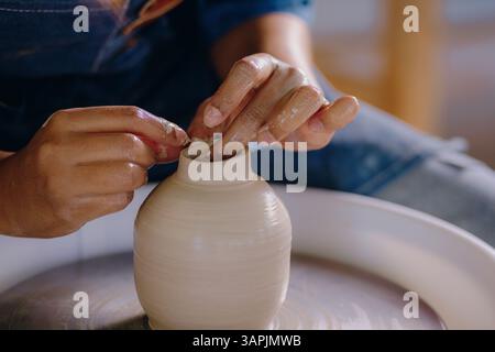 Mexikanerin, 24, formt ein Keramikstück in ihrem Atelier, trägt eine blaue Schürze und Denim. Nahaufnahme ihrer Hände, Werkzeuge und fertigen Arbeiten sichtbar. Stockfoto