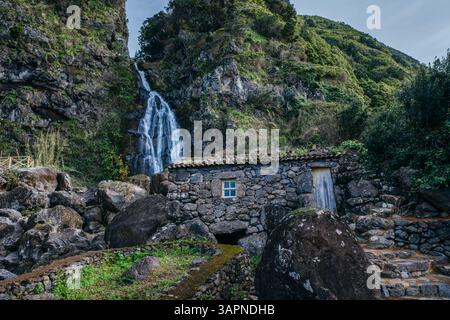 Bezauberndes Steinhaus am kaskadierenden Wasserfall: Historische Ruine in Sao Jorge, Azoren, Portugal. Rustikaler Charme inmitten natürlicher Schönheit und üppiger Landschaft Stockfoto