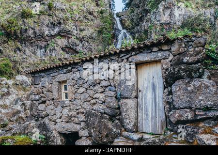 Eine charmante Ruine aus Stein liegt eingebettet zwischen Felsen in der Nähe eines wunderschönen Wasserfalls in Sao Jorge, Azoren, Portugal. Eine malerische Szene von rus Stockfoto