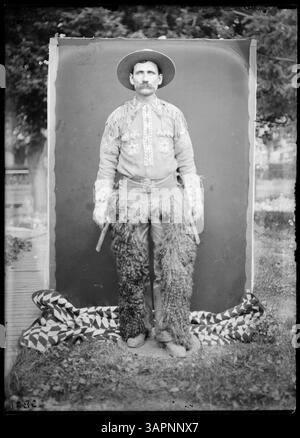 Dieses Foto zeigt Munshang, einen Cowboy im vollen Kostüm, der ein Gewehr hält. Das Porträt ist in voller Länge und zeigt die traditionelle Cowboykleidung und das Verhalten. Das Foto ist Teil der Lee Moorhouse-Sammlung der University of Oregon Libraries. Stockfoto