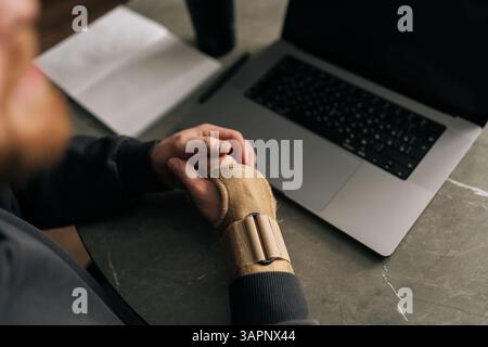 Nahaufnahme von oben auf männliche Hände, die eine Handgelenkstütze tragen, massieren schmerzhaftes Handgelenk, während sie von einem Büro aus arbeiten, umgeben von Laptop und Stockfoto