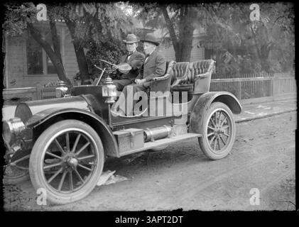 Foto von Lee Moorhouse von einer Autoparade in Pendleton, Oregon. Das Bild zeigt eine Nahaufnahme von zwei Männern in einem Auto, mit einem Haus im Hintergrund, das die Automobilkultur des frühen 20. Jahrhunderts zeigt. Stockfoto