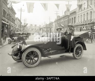 Ein Schwarzweiß-Foto von Walter S. Bowman zeigt einen Mann und eine Frau in Western-Stil-Outfits, die in einem Auto in Pendleton, Oregon, posieren. Das Auto parkt in der Mitte einer Straße, die für eine Parade dekoriert ist, mit Bannern und Fahnen sichtbar. In der Nähe säumen Unternehmen und geparkte Fahrzeuge die Straße. Stockfoto