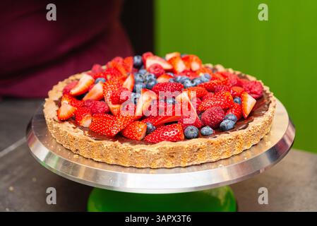 Frischer Obstkuchen mit Schokoladenfüllung und Nusskruste, belegt mit Erdbeeren, Himbeeren und Heidelbeeren auf einem Servierstand. Stockfoto