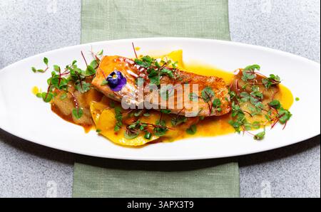 Nahaufnahme eines gebratenen Lachsfilet auf Mangoblättern mit Soße, Mikrogrün und essbarer Blüte auf weißem Teller. Stockfoto