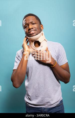 Verletzte männliche Person mit einem Halsband in einem Studio, um sich von einem Wirbelbruch zu erholen, sichtbar verärgert von Schmerzen. Junger schwarzer Mann, der unwohl mit den Händen auf der Halsbandage steht. Stockfoto