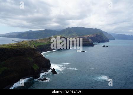 Dramatischer Blick aus der Luft auf die östlichen Klippen von Madeira, Portugal, mit Windturbinen auf dem Bergkamm, zerklüfteter Küste und türkisfarbenem Wasser. Stockfoto