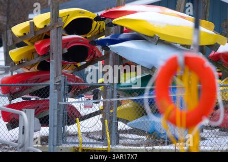 Eine lebhafte Ansammlung roter, gelber, blauer und grüner Kajaks ruhen in Regalen hinter einem umzäunten Hof, bedeckt mit frischem Schnee. Gefangen in Kingston, diese Szene Stockfoto