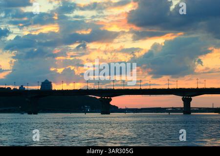 Kiew, Ukraine. 1. Mai 2021 Eine Brückensilhouette erstreckt sich über den ruhigen Fluss unter einem lebendigen Sonnenunterganghimmel, bunten Wolken, Licht reflektiert auf dem Wasser. Stockfoto