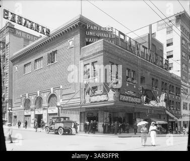 Dieses freimütige Porträt fängt das Stadtleben in Portland, Oregon, ein großes Backsteingebäude, das als Pantages Vaudeville Theater bezeichnet wird, ein. Die Oberfläche des Theaters ist mit beleuchteten Schildern geschmückt. Eine Menschenmenge versammelt sich in der Nähe des Eingangs, während sich ein Mann auf ein geparktes Auto lehnt. Zwei Frauen, eine mit einem Schirm, gehen über die Straße. Das Gebäude auf der rechten Seite ist mit "Morgan Building" gekennzeichnet. Weitere Elemente sind Straßenlaternen, elektrische Leitungen, Topfblumen und eine Treppe. Stockfoto
