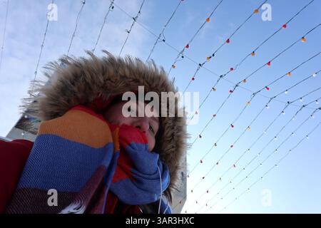 Frau posiert für ein Selbstporträt in einem roten Parka mit Kapuzenpullover, unter bunten Glühbirnen während des Winters. Blauer Himmel Stockfoto