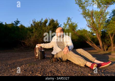 Hispanischer Reisender, der bei Sonnenaufgang am Ufer eines Baches sitzt, entspannt und die ersten Sonnenstrahlen empfängt. El Palmar Nationalpark, Entre Rios, Ar Stockfoto