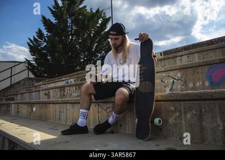 Junger sportlicher Mann, der online am Skateboard eine Nachricht liest. Stylischer Hipster auf Skate mit Mobiltelefon im Freien. Cooler Skater-Typ, der sm hält Stockfoto