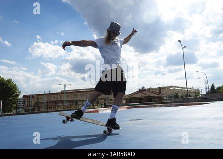Der junge blonde Hippie mit seinem Skateboard macht professionelle Tricks und Sprünge Stockfoto