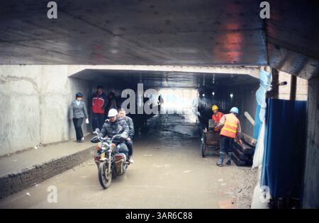 Bauarbeiter und Pendler navigieren in einem stark frequentierten unterirdischen Tunnel in einer städtischen Umgebung während der frühen Morgenstunden: China Anfang der 2000er Jahre Stadtunterführung mit Motorradverkehr Stockfoto