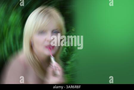 Grüner Hintergrund mit einer verschwommenen blonden, Reifen Frau, die Lippenstift-Creme auf ihre Lippen aufträgt Stockfoto