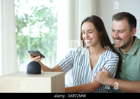Glückliches Paar, das zu Hause auf einer Couch sitzt und ein Telefon mit intelligentem Lautsprecher koppelt Stockfoto