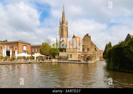 Berühmte Aussicht auf einen Kanal von Brügge mit dem Turm der Marienkirche, die über historischen Gebäuden thront. Reflektionen schimmern auf dem Wasser. Stockfoto